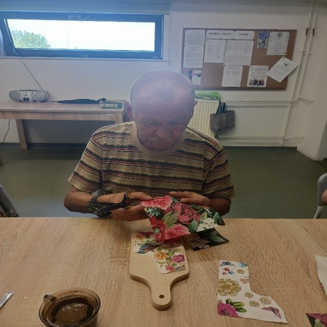 mężczyzna w pomieszczeniu; siedzi przy stole, wycina róże z serwetki, układa je na desce do krojenia