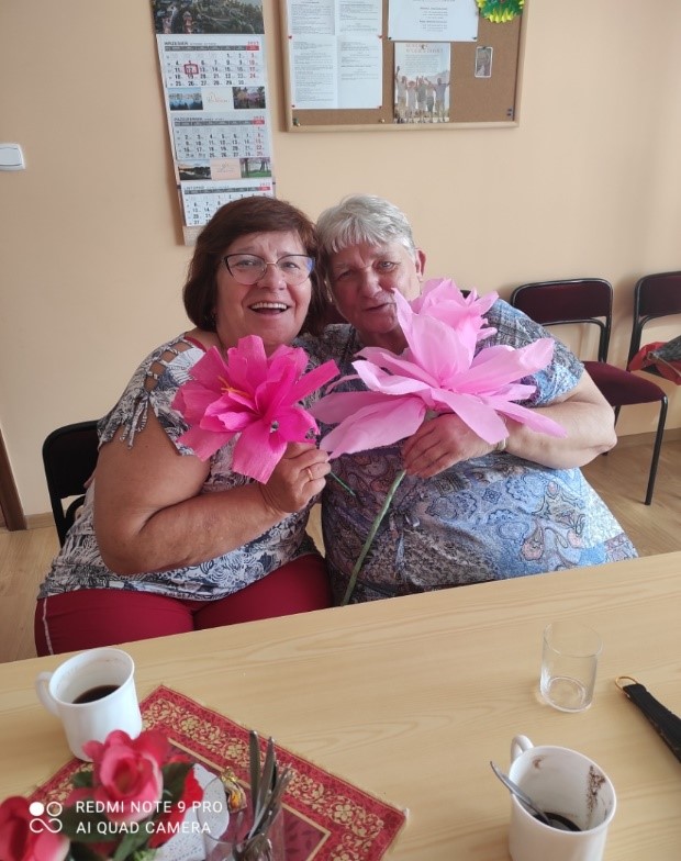 dwie wesołe kobiety seniorki, w dłoni mają różowe kwiaty z bibuły