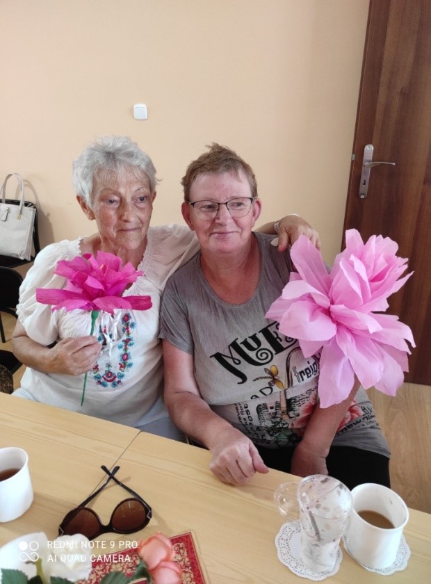 dwie kobiety, siedzą przy stole, w dłoni mają różowe kwiaty z bibuły