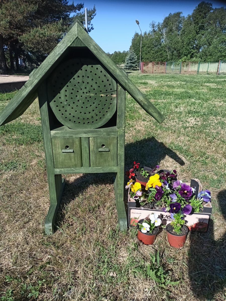 na zdjęciu widać domek dla owadów i obok kwiaty w doniczkach stojące na trawie