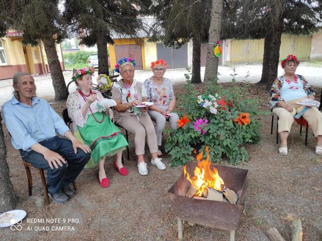 mężczyzna i cztery kobiety w wiankach na głowie siedzących przy ognisku, w tle zabudowania i drzewa